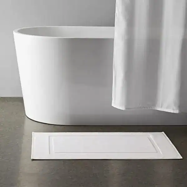 Weißer Badezimmerteppich aus Baumwolle mit Bordüre vor freistehender Badewanne.